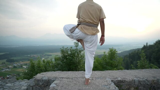 Indian man doing hatha yoga standing at the edge of a stone castle wall in the morning sun at sunrise overlooking the valley bellow with fields and woods