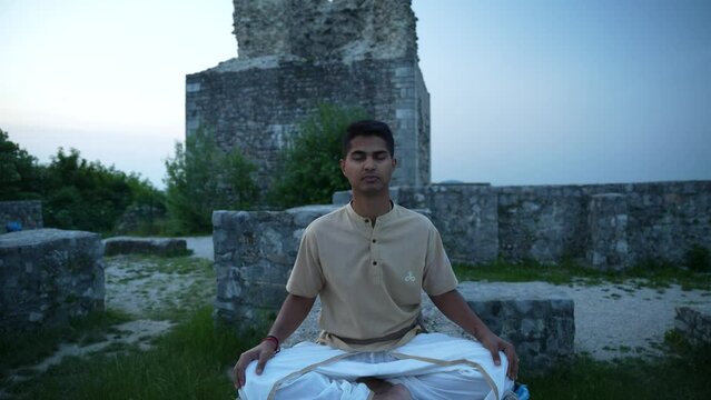 young Indian man opening his chakras with deep meditation and hatha yoga while sitting on stone wall of castle ruins on top of the hill at sunrise