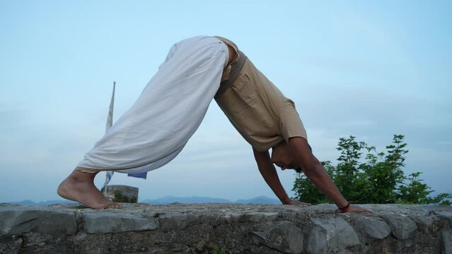 Indian man practicing his hatha yoga pose on top of a stone wall at dawn