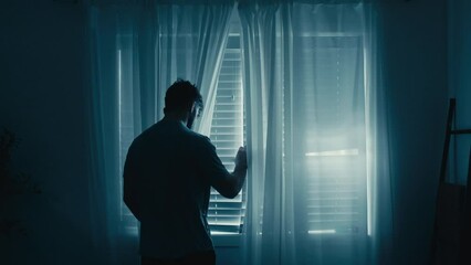Silhouette of man peering looking through blinds outside, moody blue vibe