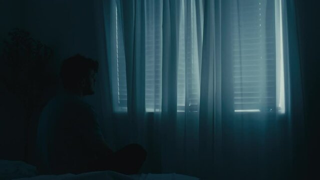 Man sitting next to bed staring out at closed window blind shades, scratching head.