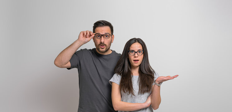 Portrait of worried and scared young couple dressed in casuals looking at camera shockingly. Amazed woman gesturing and man holding eyeglasses while standing against background