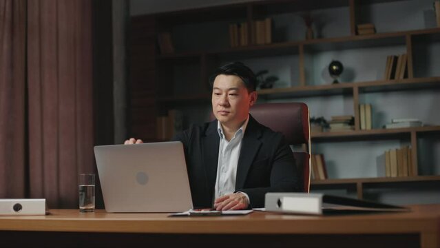 Successful entrepreneur sitting at long wooden desktop in cozy office and turning off portable laptop. Ambitious asian boss finishing workday and thinking about achievements.
