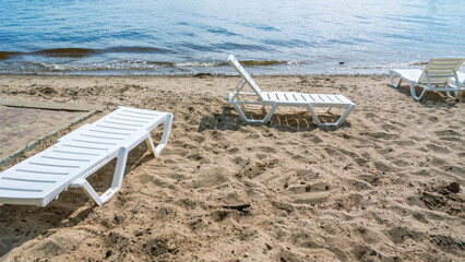Fototapeta na wymiar A beach by the river with plastic sun loungers. Sandy beach on the shore . Beach holidays in summer. White plastic sun lounger for sunbathing. Vacation at sea.