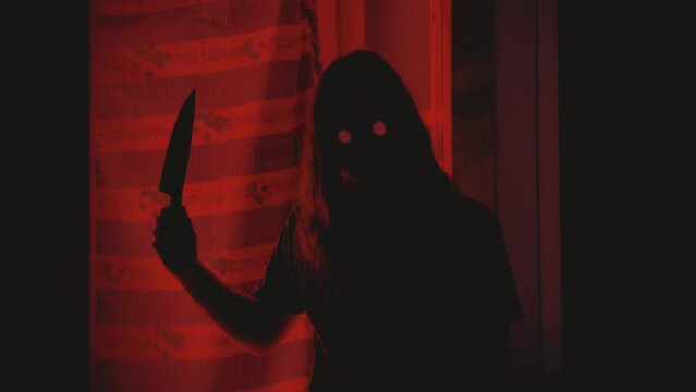 Zombie ghost girl with glowing eyes holding a big knife. Turning head toward camera. Backlit red light. Silhouette. Horror video clip. Retro vintage footage. Handheld POV camera. Halloween theme.