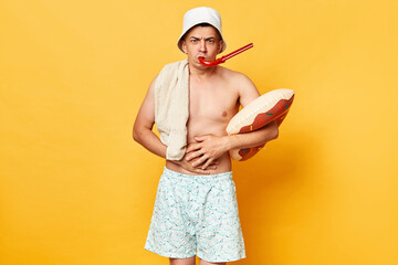 Unhappy unhealthy man wearing shorts swimsuit and panama holding donut rubber ring isolated on yellow background suffering stomachache tropical food poisoning.