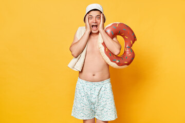Surprised overjoyed man wearing shorts swimsuit and panama holding donut rubber ring isolated on...