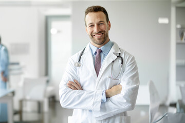 Happy healthcare worker. Portrait of confident doctor in white uniform with stethoscope posing with folded arms smiling at camera at medical clinic