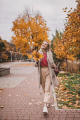 Attractive middle age woman in trendy coat in front of autumn leaves enjoys warm weather. Mature woman walks in the autumn park and enjoys an active life vertical full lenght