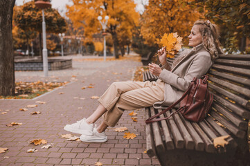 Attractive middle age woman in trendy coat in front of autumn leaves enjoys warm weather. Mature woman walks in the autumn park and enjoys an active life. Sits on a bench