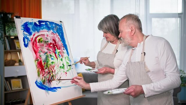leisure time in retirement, loving elderly couple have fun painting on canvas with paints and brushes using easel at home