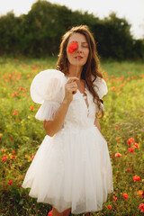 Fototapeta na wymiar Stylish brunette bride in short white wedding dress posing at the red poppies field with closed eyes and a poppy at one eye 