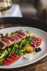 Nicoise salad with tuna, asparagus, tomatoes and a glass of white wine on a large white plate.