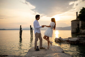 Fototapeta premium Romantic just married couple dancing looking to each other at a medieval stone pier of Italian village at Lago di Garda at the golden sunset