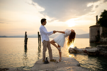 Romantic just married couple dancing looking to each other  at a medieval stone pier of Italian village at Lago di Garda at the golden sunset