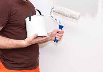 The male hand of the painter, holds a paint roller and a can of paint on the background of the wall.