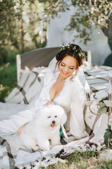 young elegant bride in a white dress with a tiara of fresh flowers on her head posing in the garden with her poodle dog