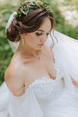 Fototapeta na wymiar a beautiful bride in a white dress with a wreath of fresh flowers is sitting on the green grass