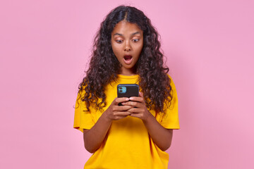 Young surprised Indian woman teenager holding phone and opening mouth wide reading shocking message about start of sale with big discounts on purchases in stands in pink studio online store.