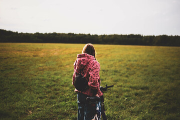 A girl wit a bicycle bicycle in a field, view from begind