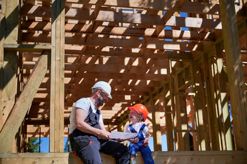 Obraz na płótnie Canvas Father with toddler son building wooden frame house. Boys having fun on the edge of balcony, examining the construction plan, wearing helmets and overalls on sunny day. Carpentry and family concept.