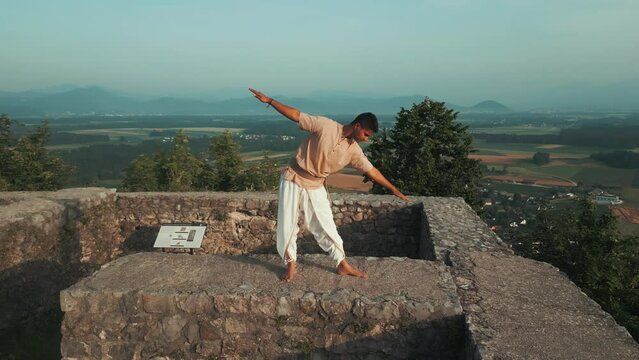 drone push-in shot of Indian man stretching and doing yoga pose on top of the hill at sunrise in traditional yogi clothes