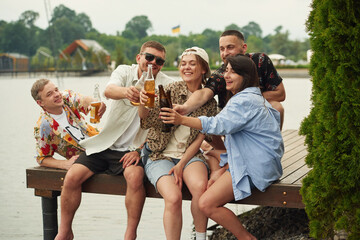 Celebrating, having fun. Group of friends are sitting near the lake with beer in hands