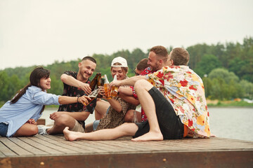 On the wooden floor. Group of friends are sitting near the lake with beer in hands