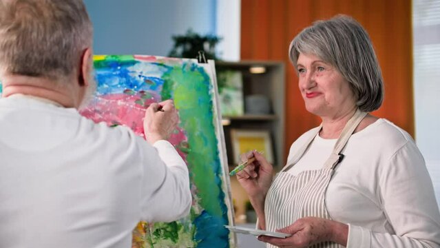 retired artists, cheerful elderly woman and man painting creative picture together using paints and brushes at home