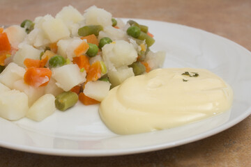 A close up view of a creamy and smooth mayonnaise sauce, served with a plate of Russian salad, a mix of potatoes, carrots, peas, tuna, and mayonnaise. A sprinkle of parsley adds some color and freshne