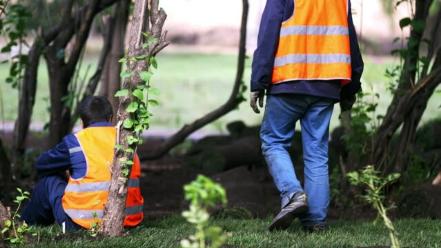 Employee walks around working place. Second man in uniform sits on grass and relaxes. City workers take break from gardening duties