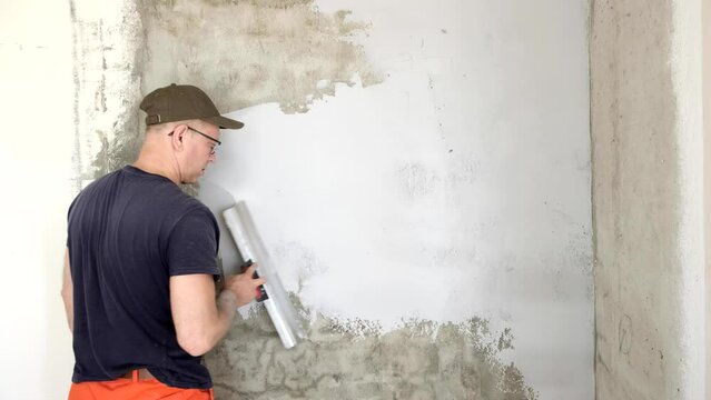 A Caucasian man, a painter, holds a spatula and puts putty on the wall.