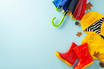 Happy autumn rain theme for kids. Creative top-down photo showcasing a colorful umbrella, yellow raincoat and rubber boots on a blue background, with copyspace for text or ads
