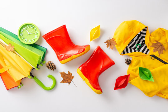 Exciting rainy day scenario in autumn for kids. Top-down image featuring green clock, rainbow umbrella, yellow raincoat, red rubber boots and colorful paper boats on a white background