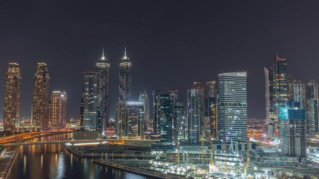 Cityscape with illuminated skyscrapers of Dubai Business Bay reflected in water canal aerial night timelapse. Modern skyline with residential and office towers on waterfront. Cranes working