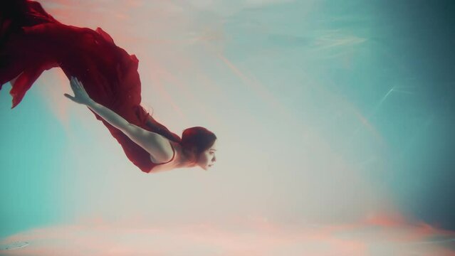 Fantasy woman mermaid swims floating in depth underwater world, long red dress fabric tail in motion. Girl princess of ocean sea blue water. magic pool creative shot. wet lady nymph siren myth goddess