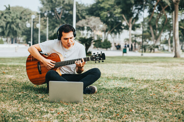 Photograph of young man playing guitar and teaching on a laptop in a park. Concept of people,...