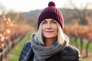 Photography in the style of pensive portraiture of a satisfied mature woman wearing a warm beanie or knit hat against a vineyard background. With generative AI technology