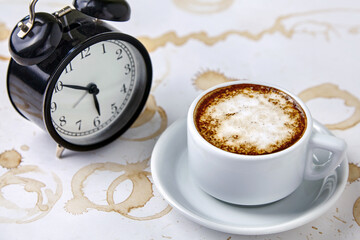 Cup of fragrant hot coffee and alarm clock on white table with coffee stains and blots. Morning cappuccino after sleepless night