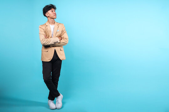 Full length portrait of a smiling young entrepreneur man standing on blue background with his legs crossed, in folded arms and looking aside to blank space 
