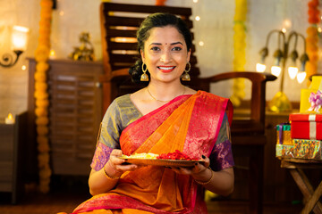 Indian happy smiling girl holding flower plate for diwali celebration by looking at camera at home...