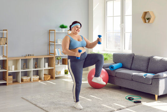 Full length photo of a happy funny young smiling fat overweight woman wearing sportswear doing fit exercise with dumbbells in living room at home. Workout sport, fitness and body positive concept.