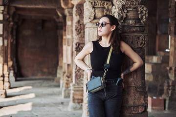 Attractive east asian woman in black sunglasses and black clothes stands among ancient pillars with ethnic patterns of old indian architecture buildings in Qutb Minar complex, full height portrait