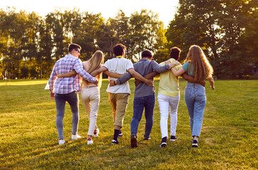 Back view of a group of happy friends standing in a row and hugging outdoors. Six young men and women wearing casual clothes spending time together. People walking in the park on a summer day.