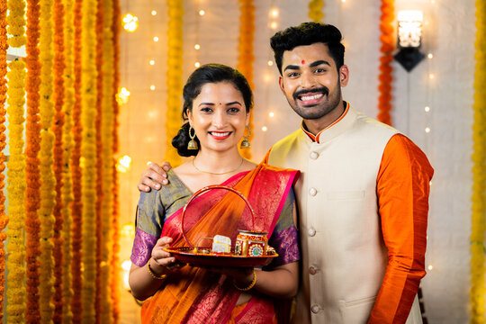 Portrait shot of happy smiling couple standing by holding karva Chauth plate set during festival celebration at home on decorated background