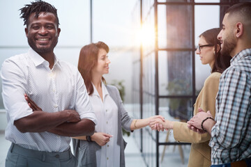 African American man posing for the camera with his arms crossed in front of his colleagues shaking hands in the office