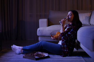 Young woman drinking from tin can and eating chips while watching TV in room at night. Bad habit