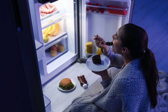 Young Woman Eating Cake Near Refrigerator In Kitchen At Night. Bad Habit