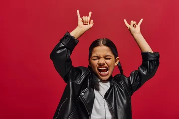 Tuinposter Excited and fashionable preteen girl with modern hairstyle closing eyes while posing in leather jacket and showing rock gesture isolated on red, girl with cool and contemporary look © LIGHTFIELD STUDIOS