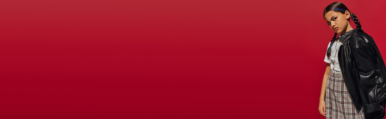 Displeased preadolescent trendy girl with hairstyle wearing leather jacket and plaid skirt while looking at camera and standing isolated on red, stylish preteen outfit concept, banner